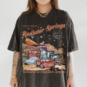 May include: Dark gray t-shirt featuring a graphic of the town of Radiator Springs from the movie Cars. The design includes the text "California Est. 2012 Radiator Springs" and "Welcome to Radiator Springs". The graphic features several cartoon cars.