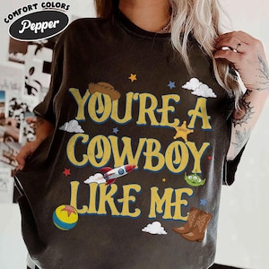 Puede incluir: Una camiseta de manga corta gris carb&oacute;n con el texto amarillo "YOU'RE A COWBOY LIKE ME" y varios elementos gr&aacute;ficos que incluyen un sombrero de vaquero, un cohete, un alien&iacute;gena verde, una pelota colorida, una estrella de sheriff y botas.