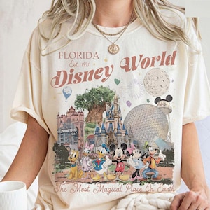 K&ouml;nnte beinhalten: Elfenbeinfarbenes T-Shirt mit einer farbenfrohen Grafik von Disney World in Florida. Das Design zeigt Wahrzeichen, Charaktere und den Text "Florida Disney World Est. 1971" und "The Most Magical Place On Earth."
