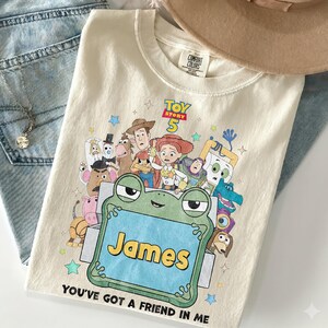 May include: A cream-colored t-shirt featuring a graphic print of Toy Story 5 characters surrounding a green frog-shaped frame with the name James in yellow text. The shirt includes the text Toy Story 5 and You've Got A Friend In Me.