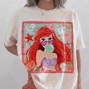Puede incluir: Camiseta de color marfil con un gr&aacute;fico colorido de una sirena con pelo rojo, gafas de sol en forma de coraz&oacute;n y una burbuja. El dise&ntilde;o incluye conchas marinas, estrellas de mar y peces. La camiseta se combina con pantalones cortos a cuadros rojos y blancos.