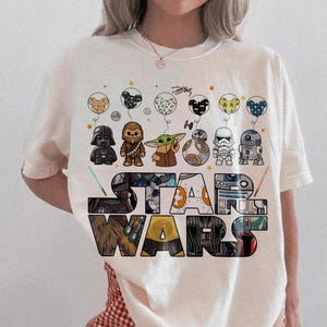 May include: Ivory t-shirt with the words "STAR WARS" in a colourful, block letter design. Above the text are illustrated balloons with Star Wars characters. The shirt has a relaxed fit.