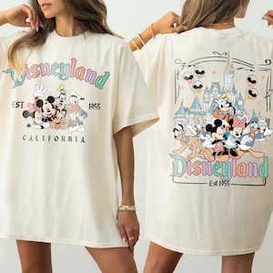 Może przedstawiać: Kremowa koszulka z logo Disneyland i postaciami z kresk&oacute;wek Myszki Miki, Myszki Minnie, Goofy'ego i Kaczora Donalda. Prz&oacute;d koszulki przedstawia napis "Disneyland Est 1955 California". Tył koszulki przedstawia zamek i logo Disneyland.