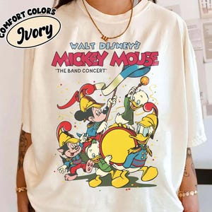 May include: Ivory-colored t-shirt featuring a graphic of Mickey Mouse and friends in a band concert scene. The text "WALT DISNEY'S MICKEY MOUSE" and "THE BAND CONCERT" are displayed in red and black. The shirt also has the words "COMFORT COLORS" and "Ivory" in a circle.