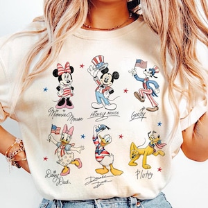 May include: Cream-coloured t-shirt featuring cartoon characters Minnie Mouse, Mickey Mouse, Goofy, Daisy Duck, Donald Duck, and Pluto. Each character is depicted in patriotic attire, holding flags or wearing festive hats. The characters' names are written below them.