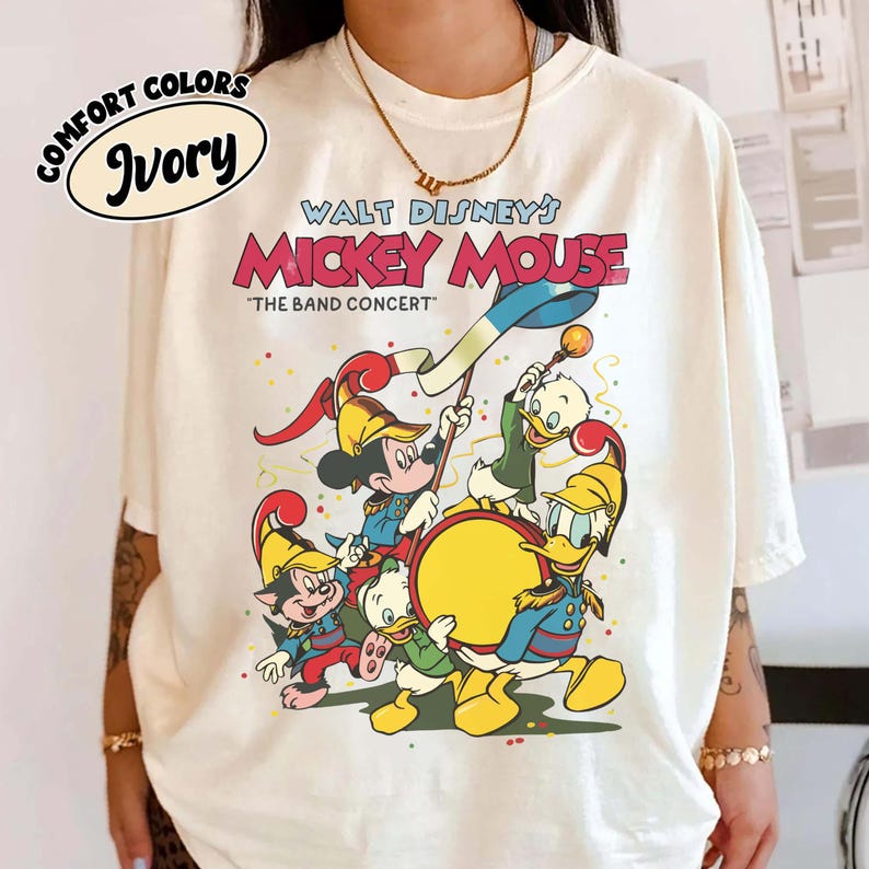 May include: Ivory-colored t-shirt featuring a graphic of Mickey Mouse and friends in a band concert scene. The text "WALT DISNEY'S MICKEY MOUSE" and "THE BAND CONCERT" are displayed in red and black. The shirt also has the words "COMFORT COLORS" and "Ivory" in a circle.