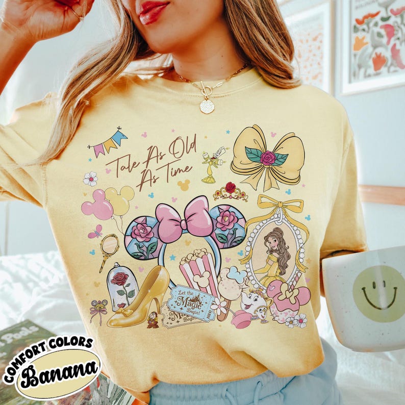 May include: Yellow t-shirt with a collage of Beauty and the Beast-themed graphics, including a portrait of Belle, a high heel, and the text "Tale As Old As Time." The shirt also features the text "Comfort Colors Banana."