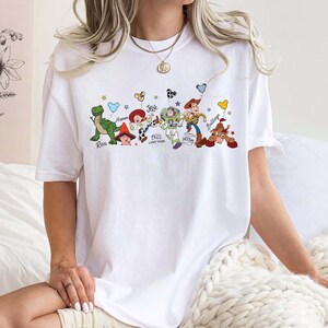 May include: White t-shirt with a vibrant graphic of Toy Story characters: Rex, Hamm, Jessie, Buzz Lightyear, Woody, and Bullseye. The design incorporates stars and balloons. The t-shirt is a relaxed, comfortable style.