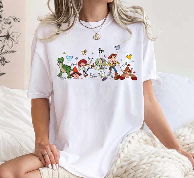 May include: White t-shirt with a vibrant graphic of Toy Story characters: Rex, Hamm, Jessie, Buzz Lightyear, Woody, and Bullseye. The design incorporates stars and balloons. The t-shirt is a relaxed, comfortable style.