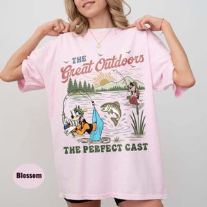 Puede incluir: Camiseta rosa claro con un dise&ntilde;o de pesca de dibujos animados con el texto "The Great Outdoors" y "The Perfect Cast". El dise&ntilde;o incluye personajes de dibujos animados, un lago, monta&ntilde;as y un sol naciente.
