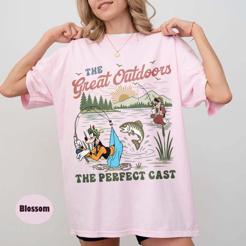 Puede incluir: Camiseta rosa claro con un dise&ntilde;o de pesca de dibujos animados con el texto "The Great Outdoors" y "The Perfect Cast". El dise&ntilde;o incluye personajes de dibujos animados, un lago, monta&ntilde;as y un sol naciente.