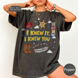 Pode incluir: Uma t-shirt cinzento-carv&atilde;o de manga curta com uma impress&atilde;o gr&aacute;fica com o texto I KNEW IT, I KNEW YOU. O design inclui um chap&eacute;u de cowboy vermelho, uma estrela de xerife dourada, blocos de letras, botas castanhas e uma pulseira.