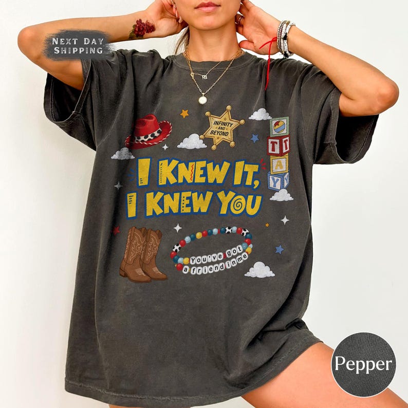 Pode incluir: Uma t-shirt cinzento-carv&atilde;o de manga curta com uma impress&atilde;o gr&aacute;fica com o texto I KNEW IT, I KNEW YOU. O design inclui um chap&eacute;u de cowboy vermelho, uma estrela de xerife dourada, blocos de letras, botas castanhas e uma pulseira.