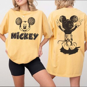 May include: Mustard yellow t-shirt with a black and white sketch of Mickey Mouse on the front and back. The front of the shirt has the word "Mickey" in black block letters. The back of the shirt shows Mickey Mouse with his arms raised in a peace sign.