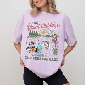 Puede incluir: Camiseta lila claro con un dise&ntilde;o de pesca con personajes de dibujos animados y el texto "The Great Outdoors" y "The Perfect Cast". La camiseta tiene un corte holgado y est&aacute; hecha de un material suave.