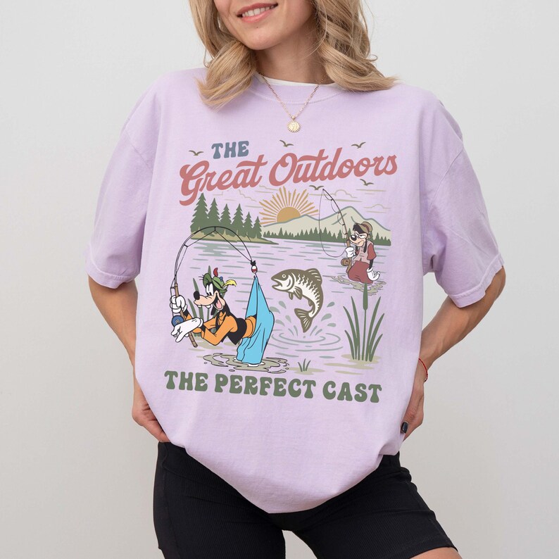 Puede incluir: Camiseta lila claro con un dise&ntilde;o de pesca con personajes de dibujos animados y el texto "The Great Outdoors" y "The Perfect Cast". La camiseta tiene un corte holgado y est&aacute; hecha de un material suave.