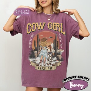 May include: A berry-colored short-sleeve t-shirt featuring a graphic print of two figures in cowboy hats standing in a desert landscape with cacti. The text reads COW GIRL and I KNEW YOU I KNEW YOU LIKE ME in a stylized font across the front.