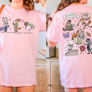 Puede incluir: Camiseta rosa claro con personajes de Zootopia. La parte delantera muestra a Judy Hopps, Nick Wilde y elementos tem&aacute;ticos de Disney con el texto "You can be anything you wish to be." La parte trasera presenta m&aacute;s personajes y texto.