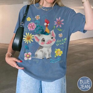 Pu&ograve; includere: T-shirt blu con un design a fumetto di maiale e gallo, circondato da fiori colorati. Il maiale &egrave; bianco con macchie grigie e un volto sorridente. Il gallo &egrave; giallo e rosso. La maglietta ha una vestibilit&agrave; ampia.