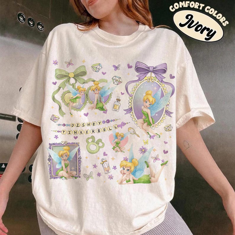 May include: An ivory t-shirt featuring multiple illustrations of Tinker Bell with green dresses and blue wings. The design includes purple bows, small perfume bottles, and the text "DISNEY TINKERBELL" in a beaded font. The top right corner displays a circular logo with the text "COMFORT COLORS IVORY".
