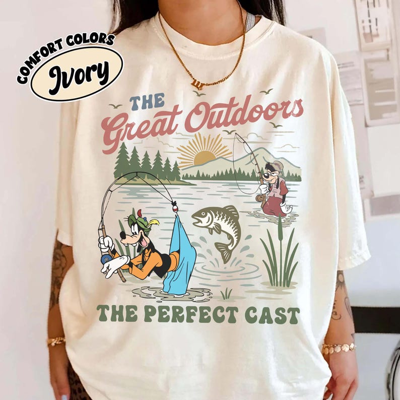 Puede incluir: Camiseta de color marfil con un gr&aacute;fico de Goofy pescando en un lago, con el texto "The Great Outdoors" y "The Perfect Cast". El dise&ntilde;o incluye un sol naciente, &aacute;rboles y un pez saltando fuera del agua. La camiseta es de la marca Comfort Colors.
