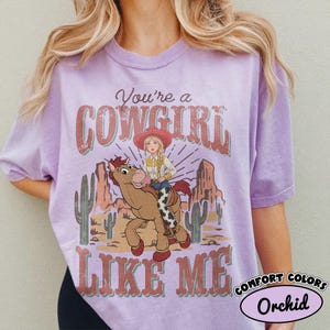 May include: A light purple short-sleeve t-shirt featuring a graphic of a cowgirl riding a brown horse in a desert landscape with cacti and rock formations. The text reads "You're a COWGIRL LIKE ME" in a distressed, vintage-style red font.