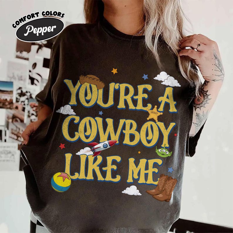 Puede incluir: Una camiseta de manga corta gris carb&oacute;n con el texto amarillo "YOU'RE A COWBOY LIKE ME" y varios elementos gr&aacute;ficos que incluyen un sombrero de vaquero, un cohete, un alien&iacute;gena verde, una pelota colorida, una estrella de sheriff y botas.