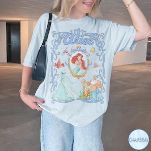 May include: Pale blue t-shirt with a framed illustration of Ariel from The Little Mermaid. The design includes Ariel's image, her dress, and King Triton. The word "Ariel" is written in a decorative font at the top of the design.
