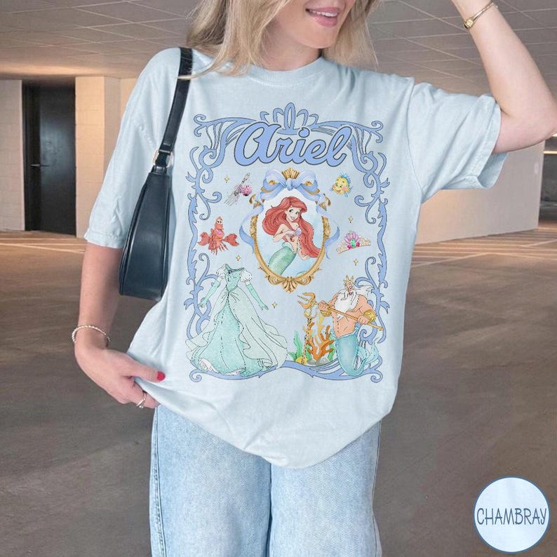 May include: Pale blue t-shirt with a framed illustration of Ariel from The Little Mermaid. The design includes Ariel's image, her dress, and King Triton. The word "Ariel" is written in a decorative font at the top of the design.