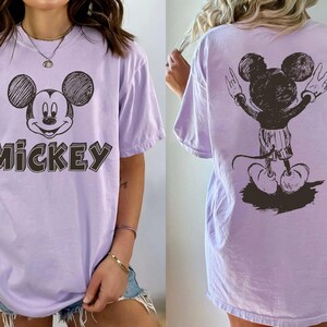 May include: A lilac t-shirt with a sketch of Mickey Mouse on the front and back. The front of the shirt has the word "Mickey" in a block letter style. The back of the shirt shows Mickey Mouse with his arms up in a peace sign.