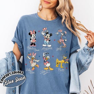 May include: Blue denim-coloured t-shirt featuring cartoon characters Mickey Mouse, Minnie Mouse, Goofy, Daisy Duck, Donald Duck, and Pluto in patriotic outfits. The shirt has the words "COMFORT COLORS" and "Blue Jean" printed on it.