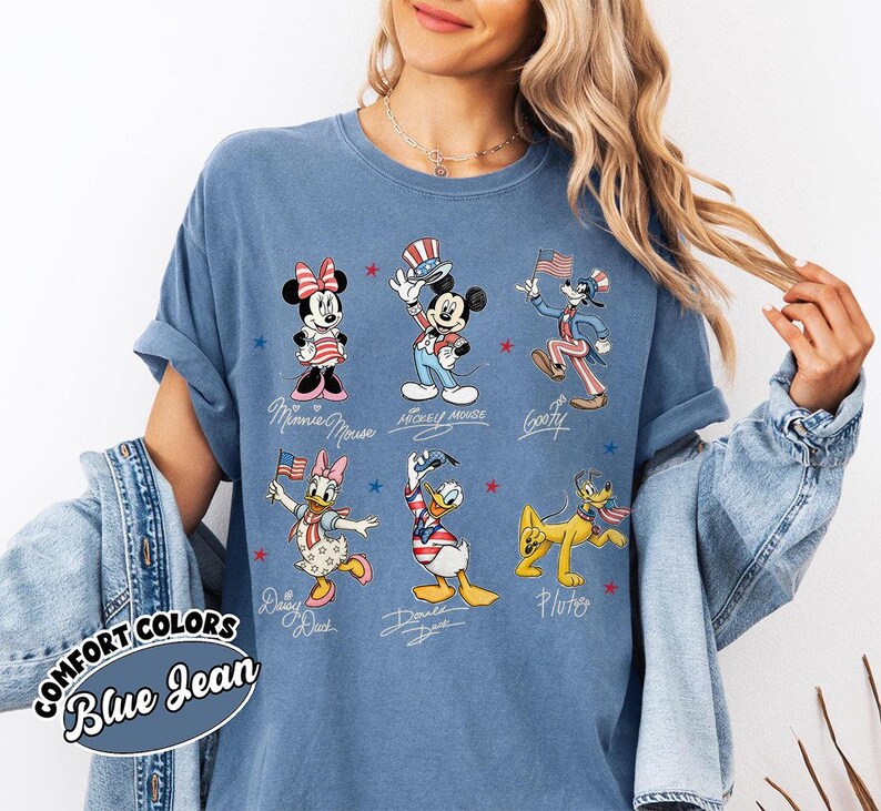 May include: Blue denim-coloured t-shirt featuring cartoon characters Mickey Mouse, Minnie Mouse, Goofy, Daisy Duck, Donald Duck, and Pluto in patriotic outfits. The shirt has the words "COMFORT COLORS" and "Blue Jean" printed on it.