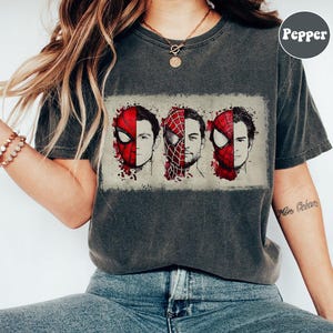May include: Dark gray t-shirt featuring a graphic of three Spider-Man faces, each with a different actor's likeness. The design includes red and black elements, with a distressed, vintage look. The shirt has short sleeves and a crew neck.
