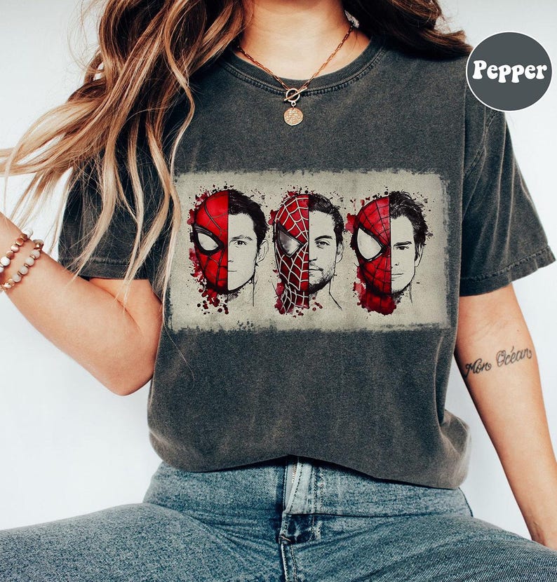 May include: Dark gray t-shirt featuring a graphic of three Spider-Man faces, each with a different actor's likeness. The design includes red and black elements, with a distressed, vintage look. The shirt has short sleeves and a crew neck.