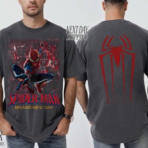 以下が含まれることがあります： スパイダーマンと都市景観を背景にした濃いグレーのTシャツ。Tシャツの前面には「SPIDER-MAN BRAND NEW DAY」の文字が赤と青で表示されています。Tシャツの背面には赤いクモのシンボルがあります。