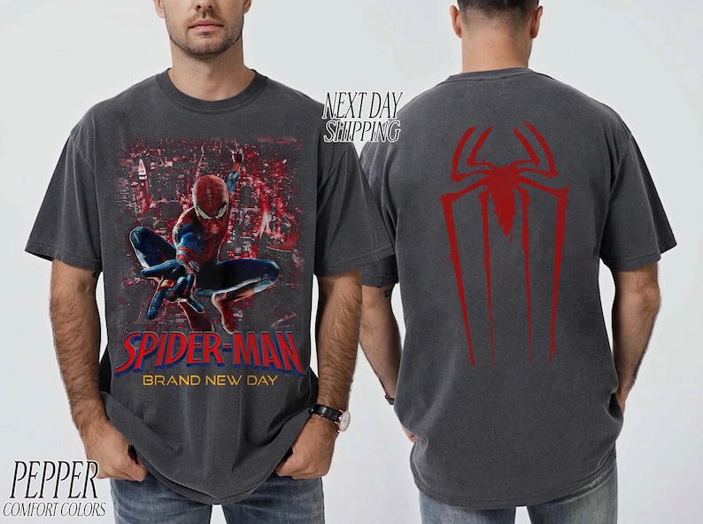 以下が含まれることがあります： スパイダーマンと都市景観を背景にした濃いグレーのTシャツ。Tシャツの前面には「SPIDER-MAN BRAND NEW DAY」の文字が赤と青で表示されています。Tシャツの背面には赤いクモのシンボルがあります。