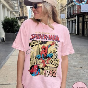 May include: A light pink t-shirt featuring a Spider-Man graphic. The design includes the words "BRAND NEW DAY", "SPIDER-MAN", "NEW YORK", and "1962". The graphic depicts Spider-Man in action with a cityscape background.