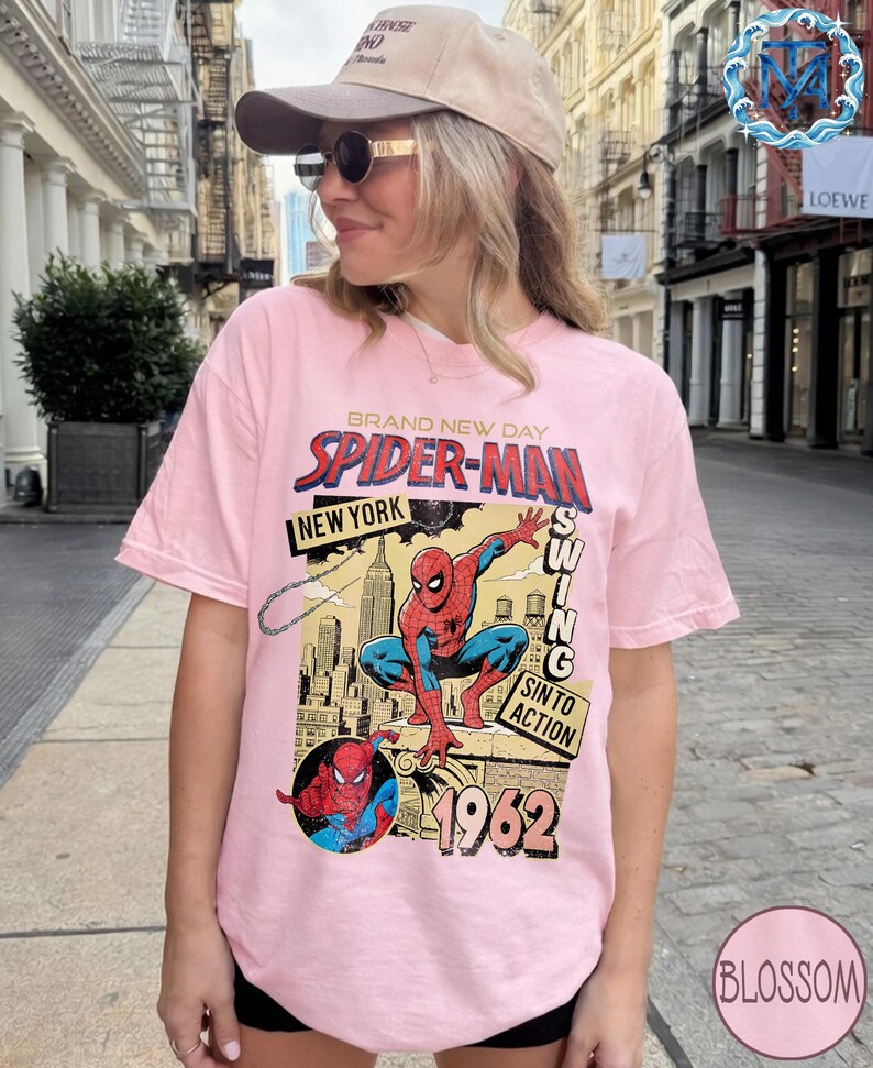 May include: A light pink t-shirt featuring a Spider-Man graphic. The design includes the words "BRAND NEW DAY", "SPIDER-MAN", "NEW YORK", and "1962". The graphic depicts Spider-Man in action with a cityscape background.