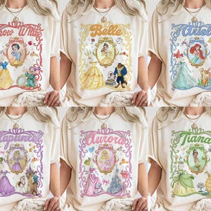 May include: Cream-coloured t-shirts with vibrant illustrations of Disney princesses. Each shirt showcases a princess's name within a decorative frame, accompanied by associated characters and imagery. The princesses featured are Snow White, Belle, Ariel, Rapunzel, Aurora, and Tiana.