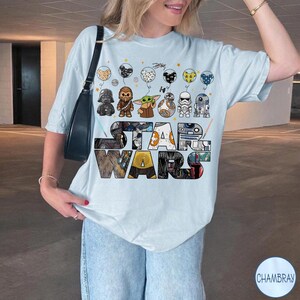 May include: Pale blue t-shirt with a multi-coloured "STAR WARS" graphic. The design includes Star Wars characters and balloons. The t-shirt is worn with blue jeans.