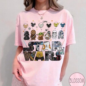 May include: Pale pink t-shirt with a Star Wars theme. The design features Star Wars characters as balloons above the colorful Star Wars logo. Characters include Darth Vader, Chewbacca, and Baby Yoda.