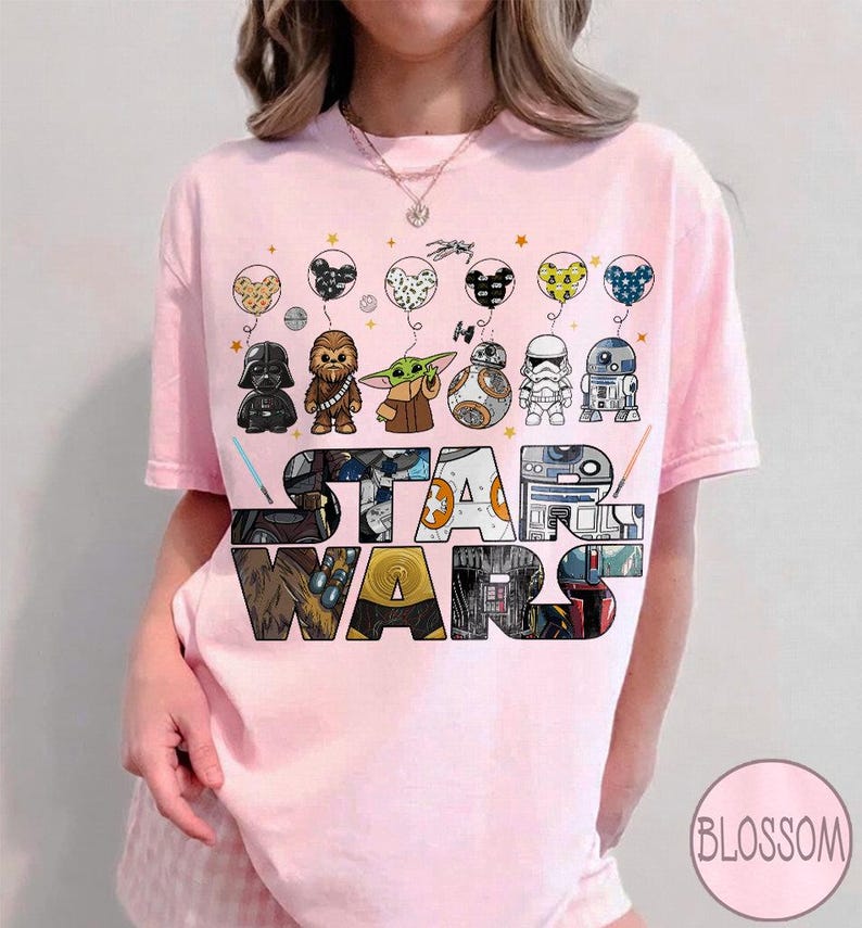 May include: Pale pink t-shirt with a Star Wars theme. The design features Star Wars characters as balloons above the colorful Star Wars logo. Characters include Darth Vader, Chewbacca, and Baby Yoda.