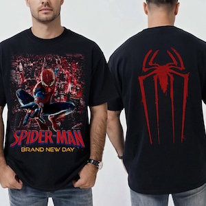 以下が含まれることがあります： スパイダーマンが都市の背景を前にダイナミックなポーズをとっている黒いTシャツ。「SPIDER-MAN BRAND NEW DAY」の文字が赤と青で表示されています。背面には赤いクモのシンボルがあります。
