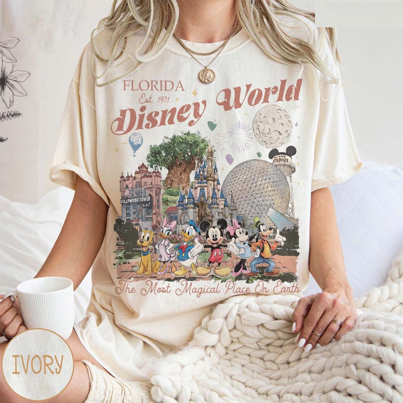 K&ouml;nnte beinhalten: Elfenbeinfarbenes T-Shirt mit einer farbenfrohen Grafik von Disney World in Florida. Das Design zeigt Wahrzeichen, Charaktere und den Text "Florida Disney World Est. 1971" und "The Most Magical Place On Earth."