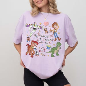 May include: A lilac t-shirt with a vibrant graphic of animated film characters. The design includes Buzz Lightyear, Woody, and other characters, along with the phrase "You've Got a Friend in Me."