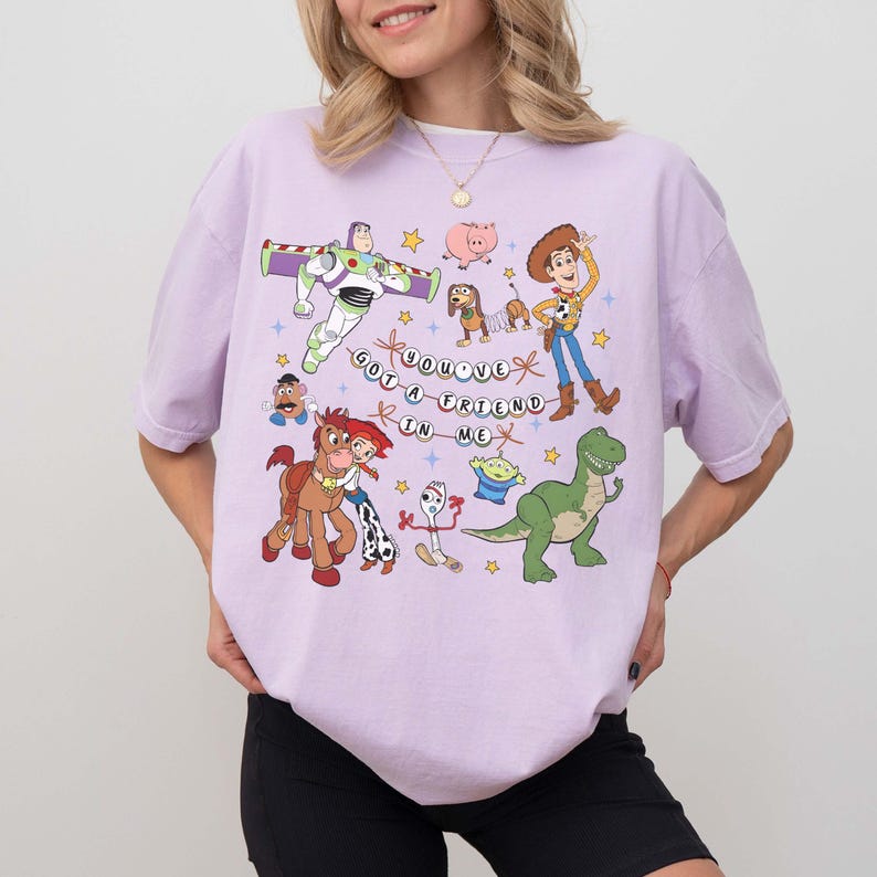 May include: A lilac t-shirt with a vibrant graphic of animated film characters. The design includes Buzz Lightyear, Woody, and other characters, along with the phrase "You've Got a Friend in Me."
