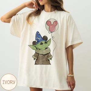 Puede incluir: Camiseta de color marfil con una ilustraci&oacute;n de dibujos animados de un personaje alien&iacute;gena verde que lleva un sombrero de mago y sostiene un globo de Mickey Mouse. El dise&ntilde;o incluye acentos marrones, azules y rosas. La palabra "IVORY" est&aacute; impresa en la parte inferior izquierda.