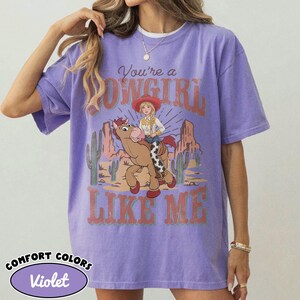 May include: A violet short-sleeve t-shirt featuring a graphic print of a cartoon cowgirl riding a brown horse in a desert landscape with cacti and rock formations. The text reads You're a COWGIRL LIKE ME in a distressed red and white font style.