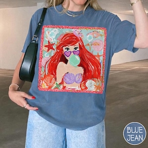 Puede incluir: Camiseta azul con un gr&aacute;fico de sirena de dibujos animados. La sirena tiene pelo rojo, gafas de sol en forma de coraz&oacute;n y una burbuja. El gr&aacute;fico est&aacute; enmarcado con un borde rosa y rodeado de conchas marinas y estrellas de mar.