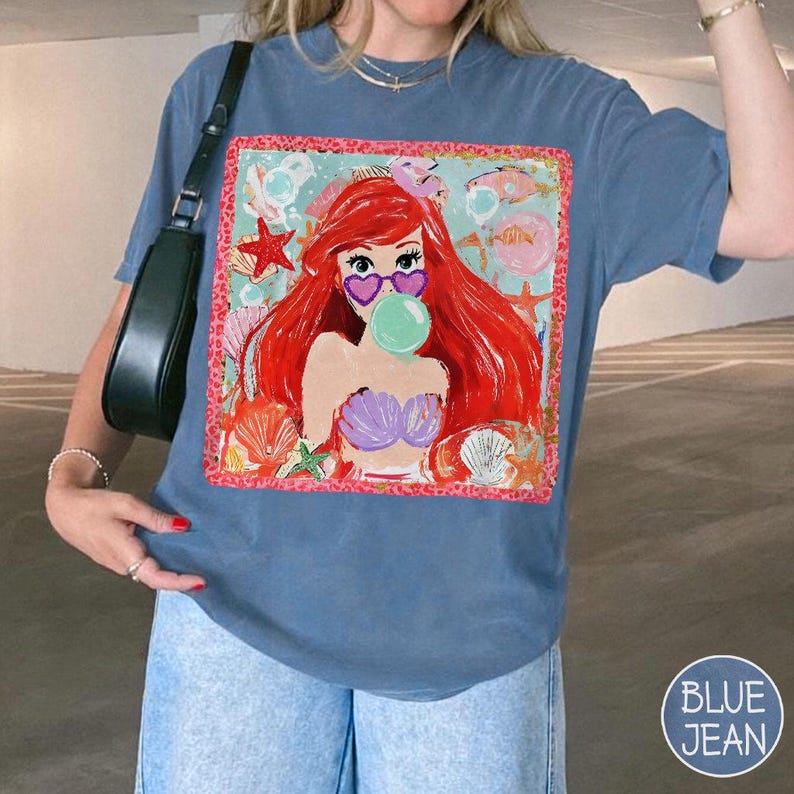 Puede incluir: Camiseta azul con un gr&aacute;fico de sirena de dibujos animados. La sirena tiene pelo rojo, gafas de sol en forma de coraz&oacute;n y una burbuja. El gr&aacute;fico est&aacute; enmarcado con un borde rosa y rodeado de conchas marinas y estrellas de mar.